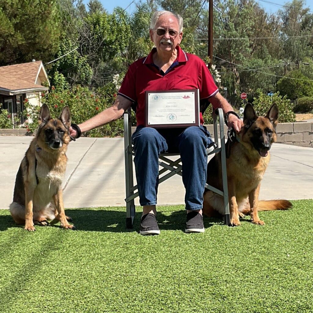Ron Cable seated on a lawn holding his certificate, with a German Shepherd on each side