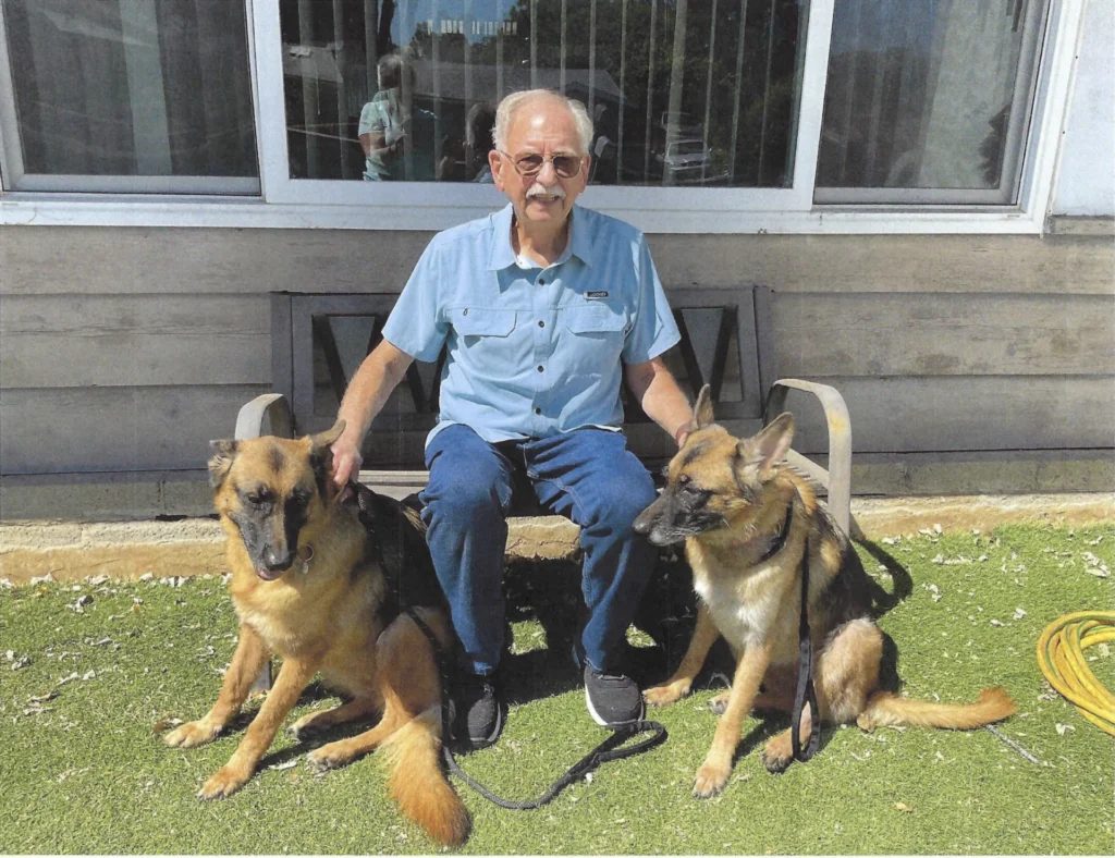 Ron Cable sitting on a bench outside with two German Shepherds at his feet
